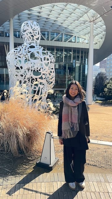 Picture of a woman wearing all black with a pink checked scarf standing in front of a building in Tokyo