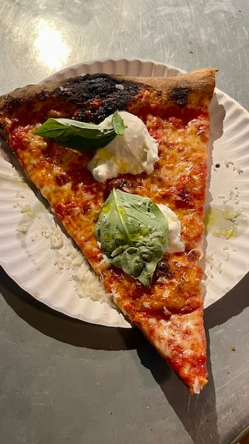 picture of a slice of burrata pizza on a paper plate on a steel counter