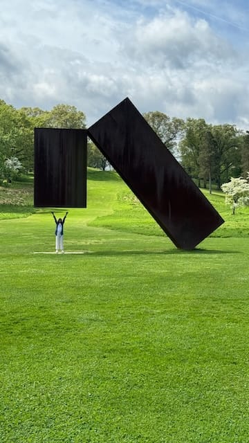 picture of a sculpture in a green grassy field and a small woman trying to lift it up