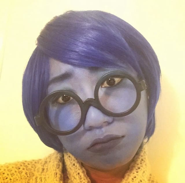 Picture of a woman dressed as Sadness. She has blue face paint, black wide-rimmed glasses, and a short blue wig.