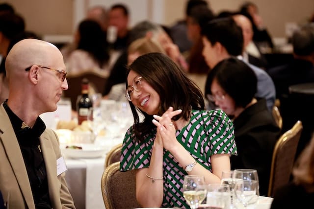 Picture of a woman laughing with a man at a table with wine glasses and people seated in the background