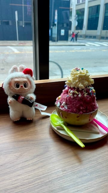 Picture of a bowl of pink-colored shaved ice with cream on top. Next to the bowl is a troll-like charm.